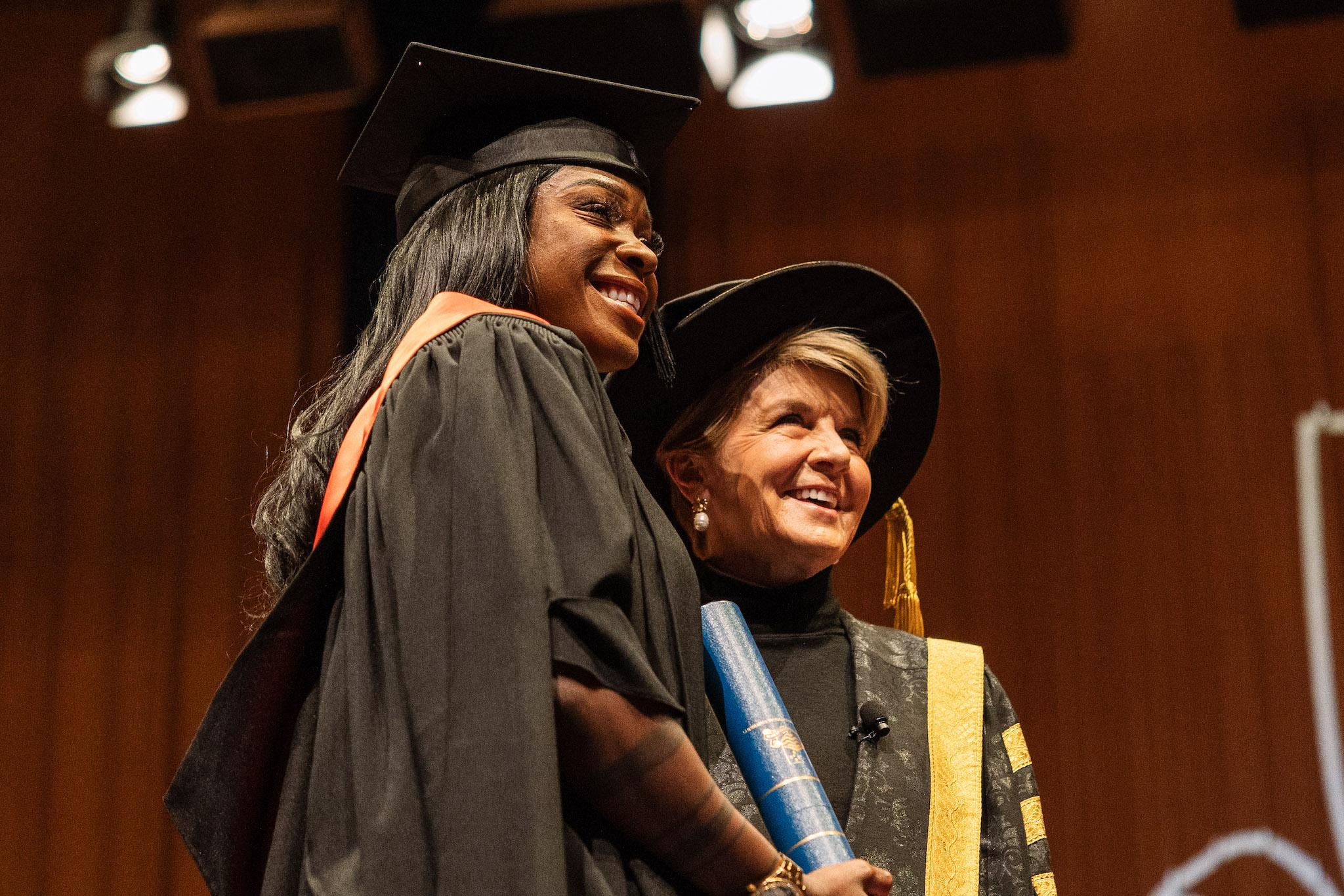 Precilla Anna-Kaii Lawrence standing on stage with Julie Bishop in graduation robes, holding her Testamur