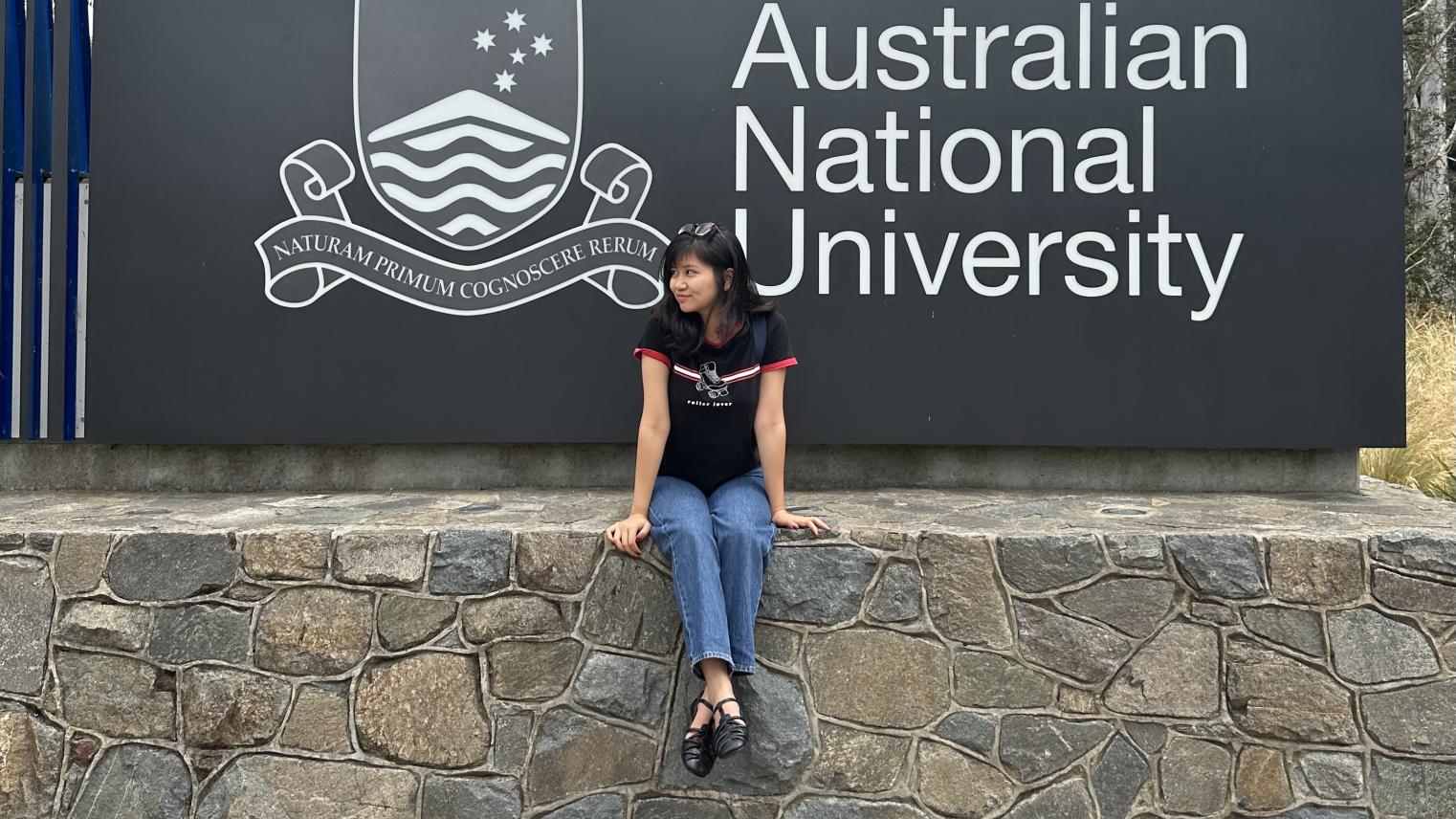 International postgraduate student Binh Thanh Ho sitting on a stone wall in front of a sign for the Australian National University