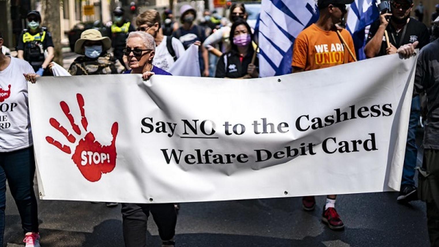 Image of banner opposing the Cashless Welfare Debit Card at a 2021 ‘Demand a Living Wage for all’ rally in Melbourne, Victoria, by Matt Hrkac on flickr https://flic.kr/p/2kMhtEh, free to use under CC BY 2.0 DEED licence 